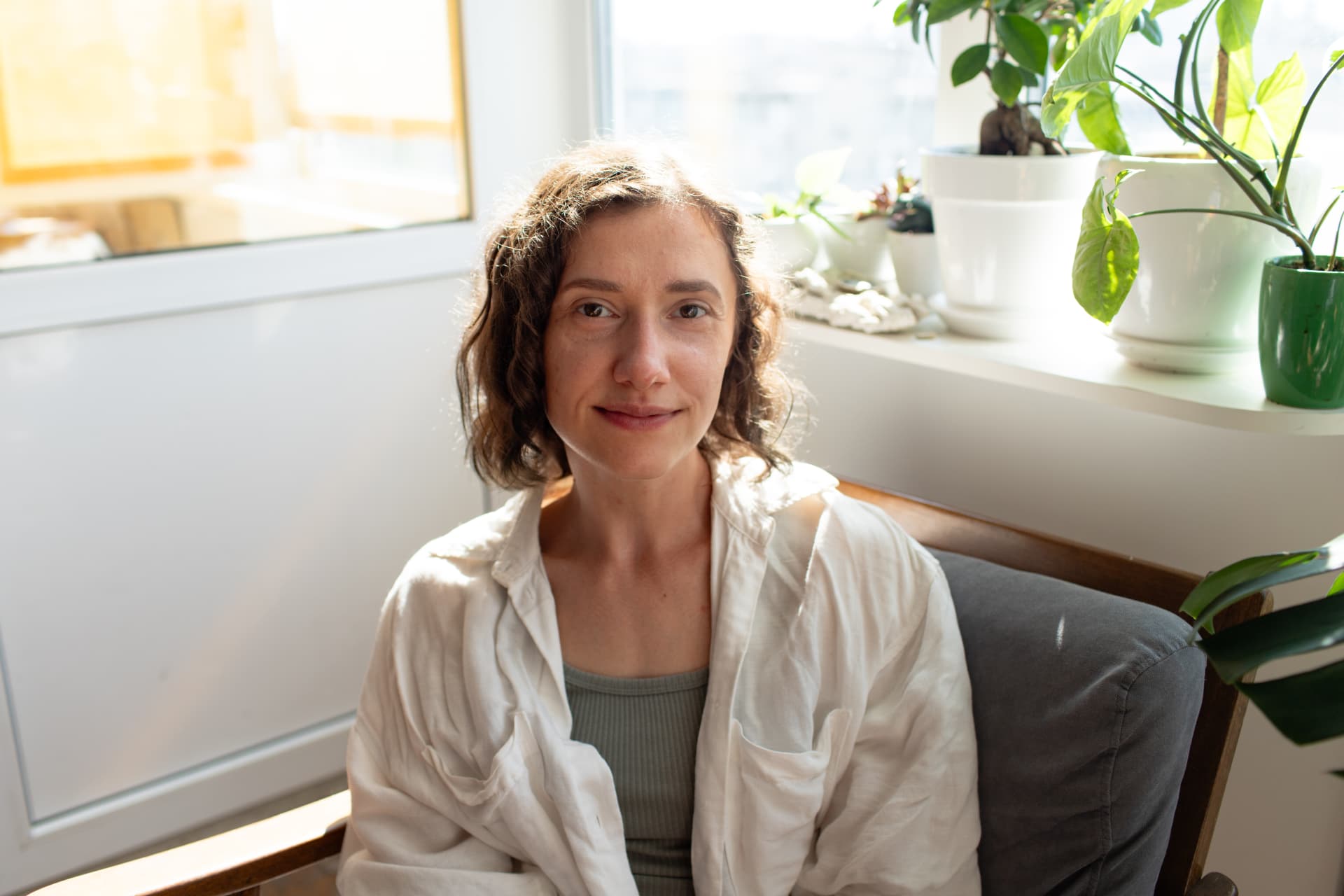 Woman with curly hair in a cream shirt, sitting by a sunny window